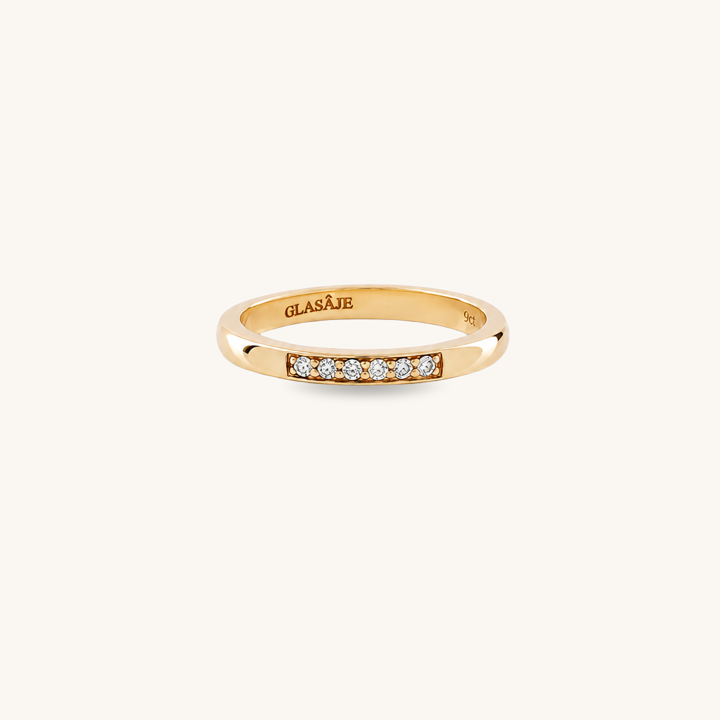 A yellow gold ring with a row of small clear gemstones on the front, shown on a plain off-white background. The inside of the band is engraved with the brand name 'GLASAJE'.