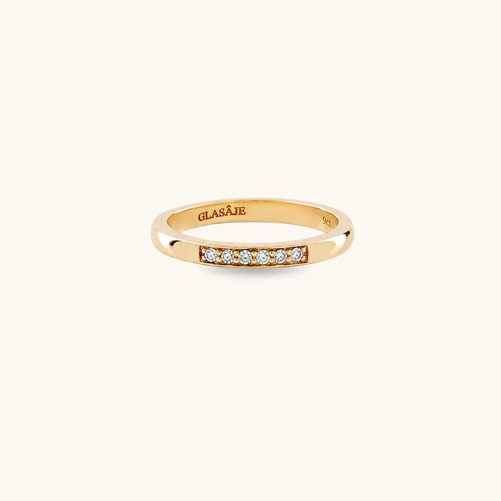 A yellow gold ring with a row of small clear gemstones on the front, shown on a plain off-white background. The inside of the band is engraved with the brand name 'GLASAJE'.