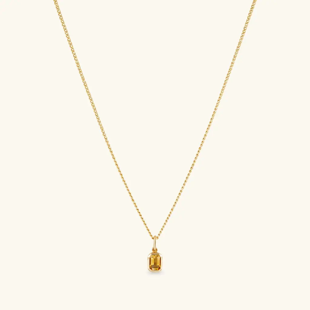 A delicate gold chain necklace with a small rectangular baguette-cut yellow gemstone pendant, displayed against a solid off-white background.