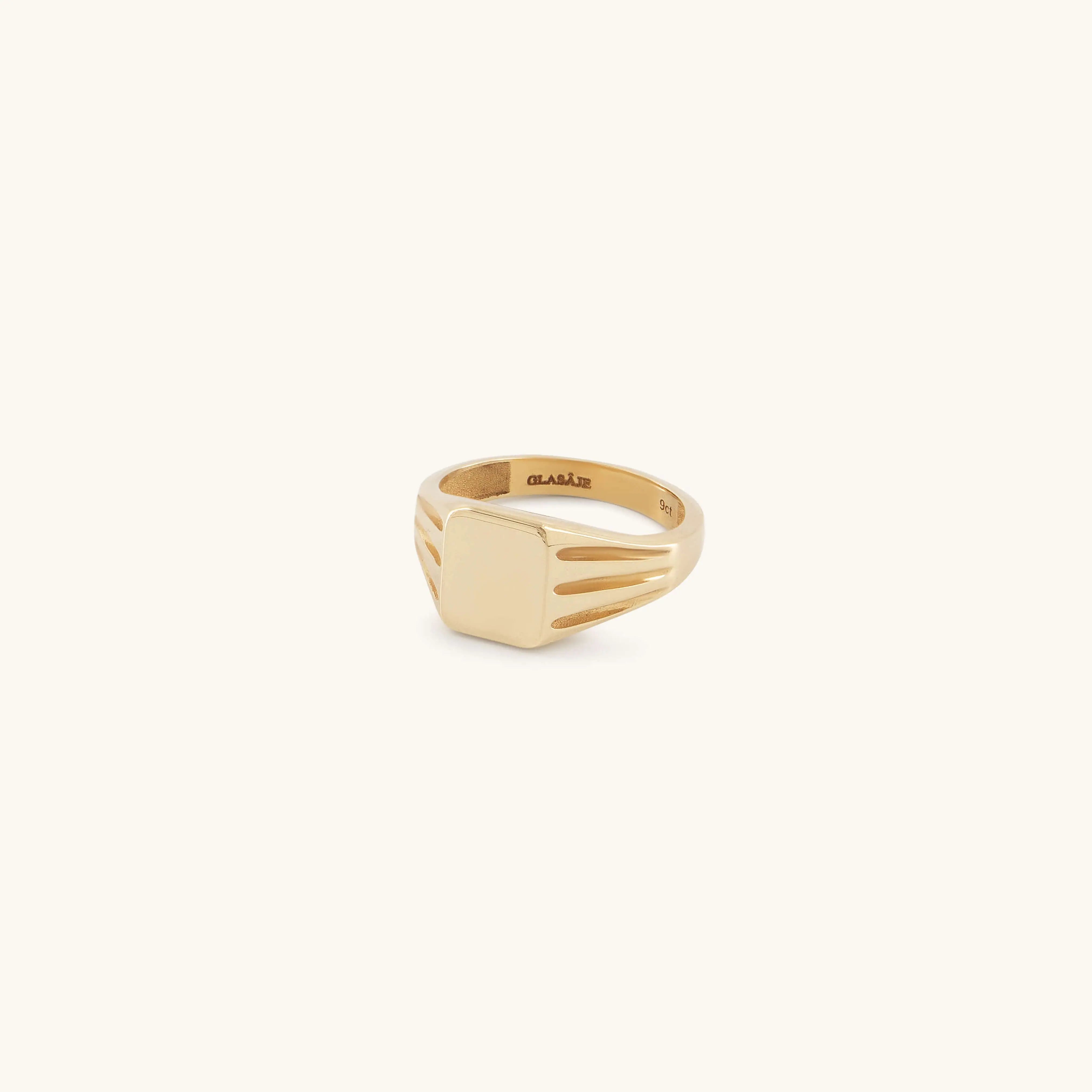 The Glasâje Signet Ring is crafted in solid gold with a rectangular flat face and debossed details, plus decorative grooves on the band, showcased against a light cream background.