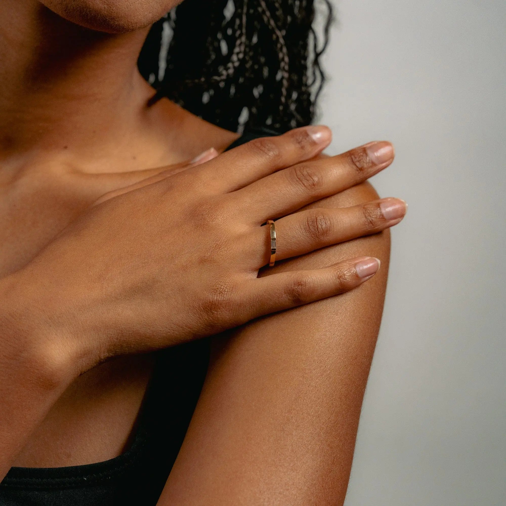 Close-up of a hand resting on a shoulder, showcasing Glasâjes Siglo Ring—an ethical diamond set in a solid gold band. The person has medium brown skin and wears a black sleeveless top against a neutral background.