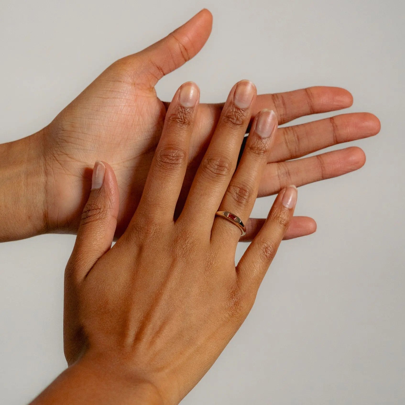 Two hands with short, natural nails are shown against a neutral background. The ring finger of one hand wears Glasâjes Tawny Ring—a solid gold signet band set with a small red stone—while the other hand is open beneath it.