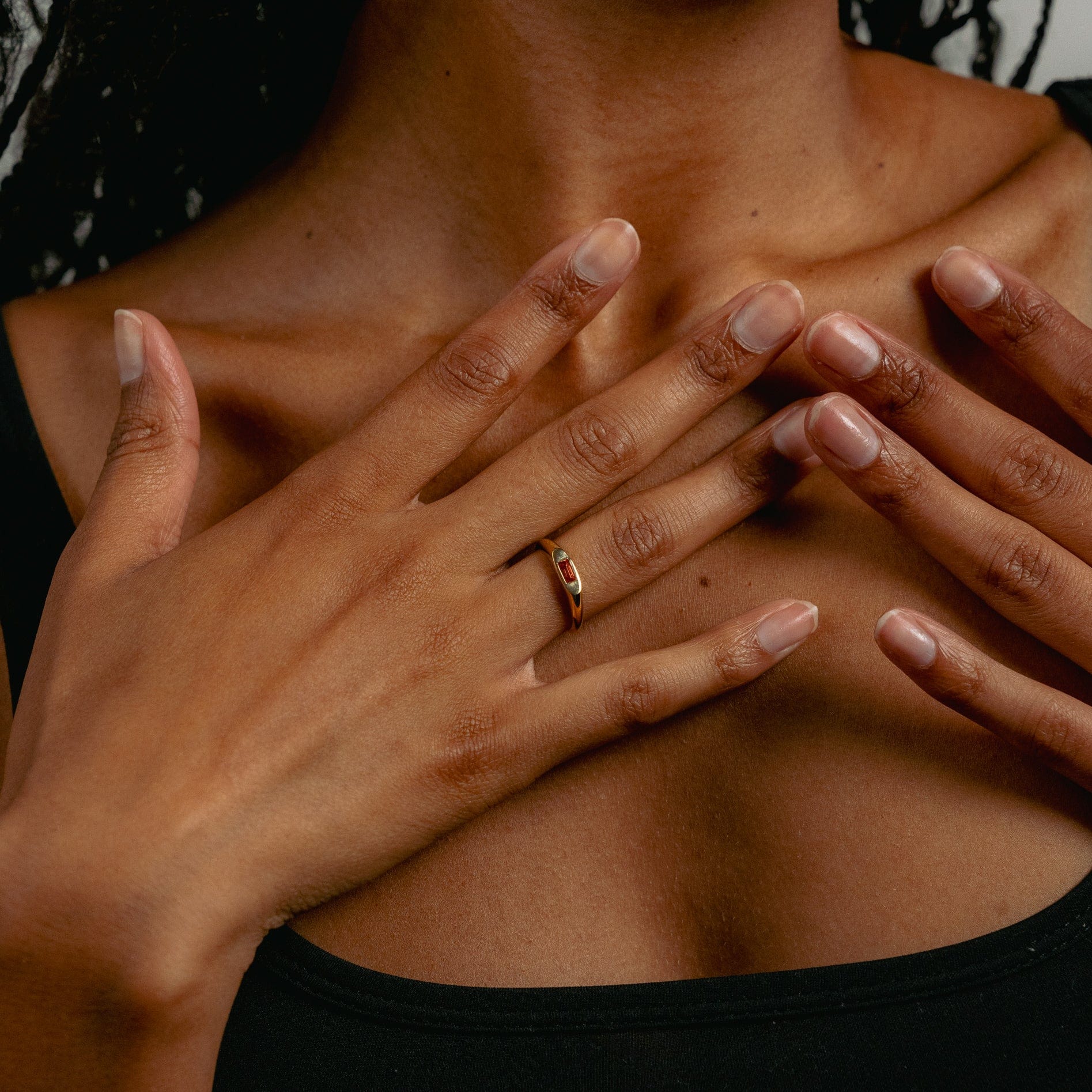 A person with dark skin wearing a black top holds their hands to their chest, showcasing the Glasâje Tawny Ring—a solid gold signet band with a small red stone—on one finger. Their nails are natural and neatly trimmed.