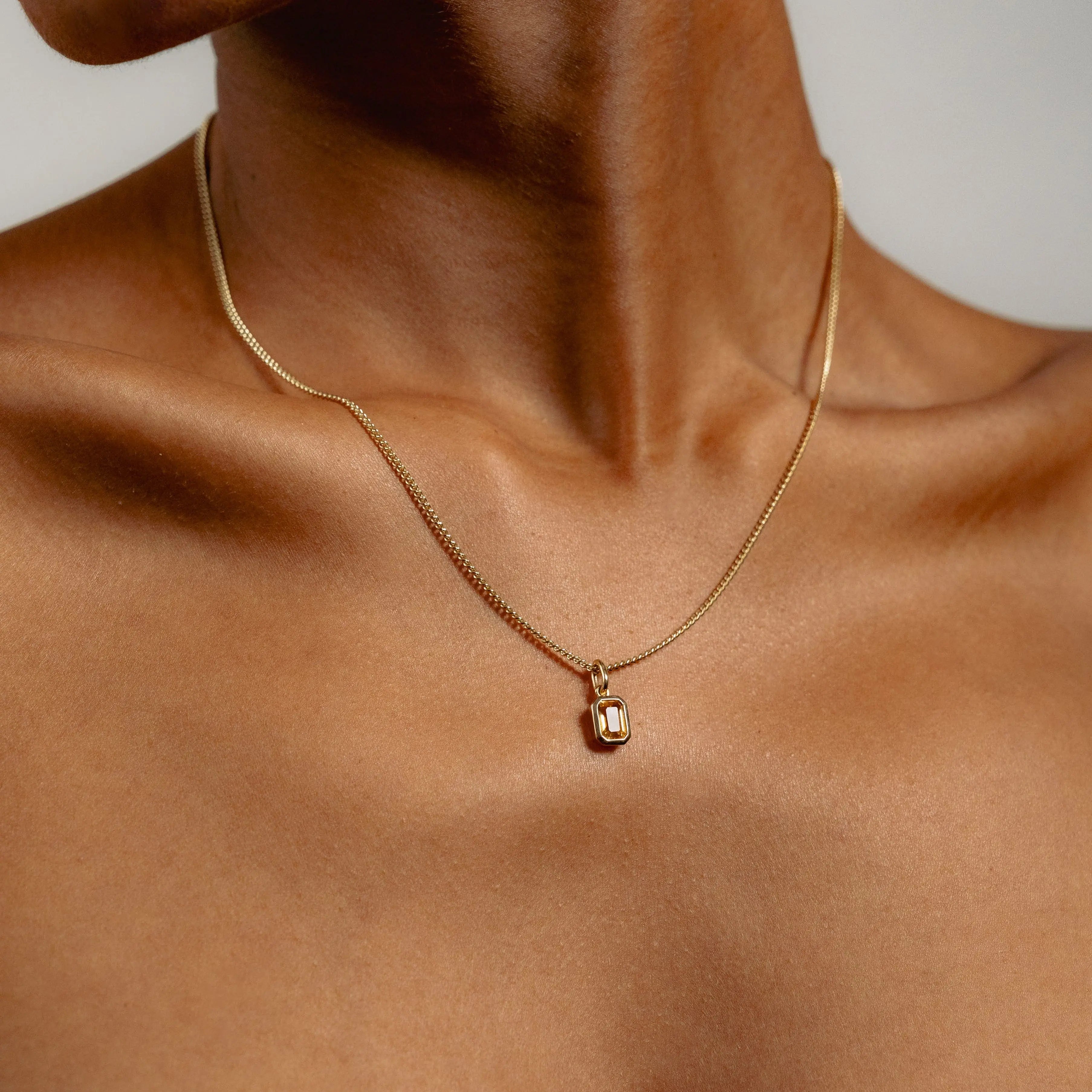Close-up of a person wearing a delicate gold necklace with a rectangular auburn citrine pendant. The skin is smooth, and the image exudes a sense of elegance and simplicity.