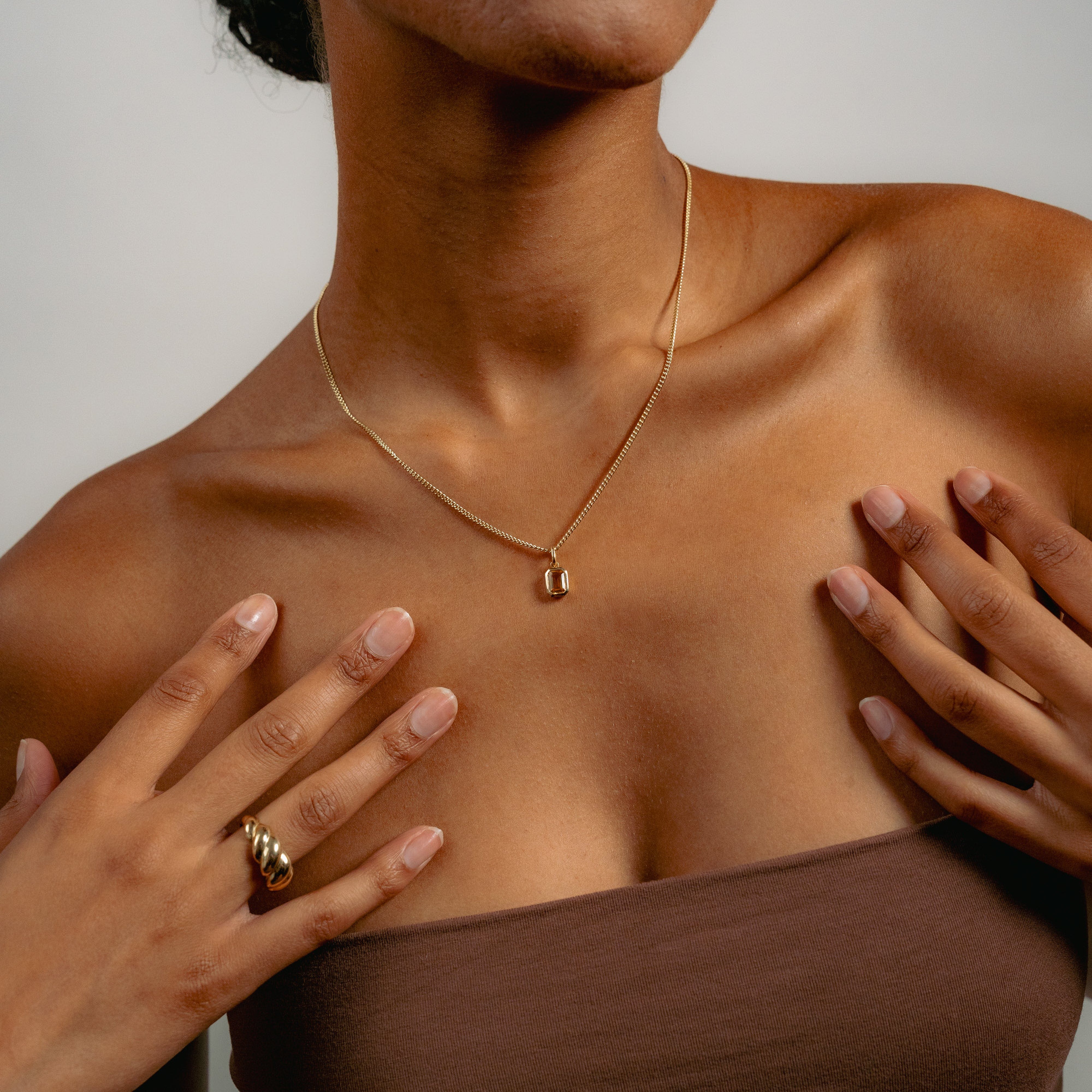 Close-up of a woman wearing a gold necklace with an auburn citrine pendant. Her hands gently touch her chest, showcasing a gold ring. She wears a strapless brown top.