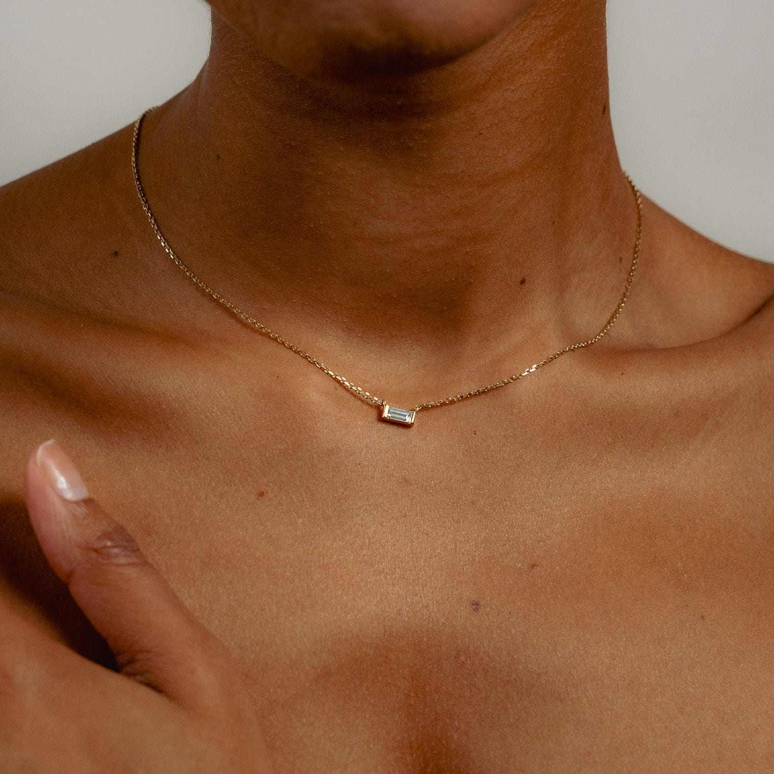A woman with medium brown skin wears a delicate gold chain necklace with a small baguette diamond pendant, shown from the shoulders up, with one hand gently touching their collarbone.