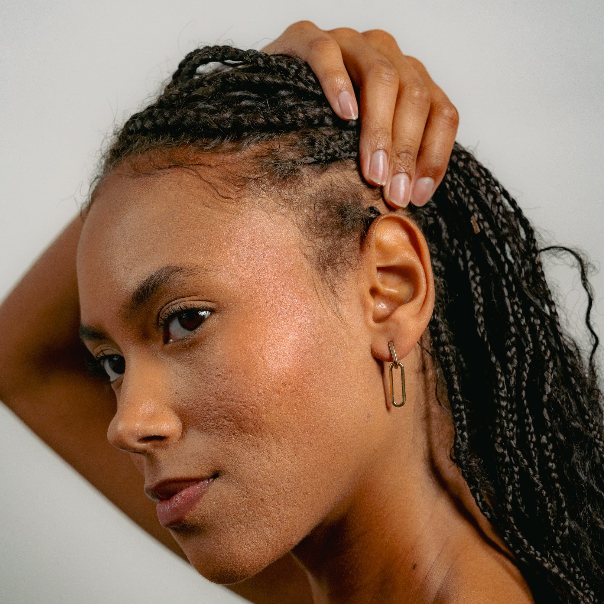 A woman with braided hair touches her head, revealing her scalp. She confidently showcases Glasâje Paperclip Earrings, highlighting their versatile style against a plain background.