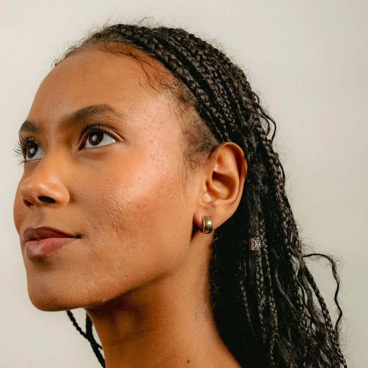 A woman with braided hair looks upward and left, wearing Glasâjes Gabriel Huggies—small solid gold earrings. A plain, light background highlights this timeless staple.