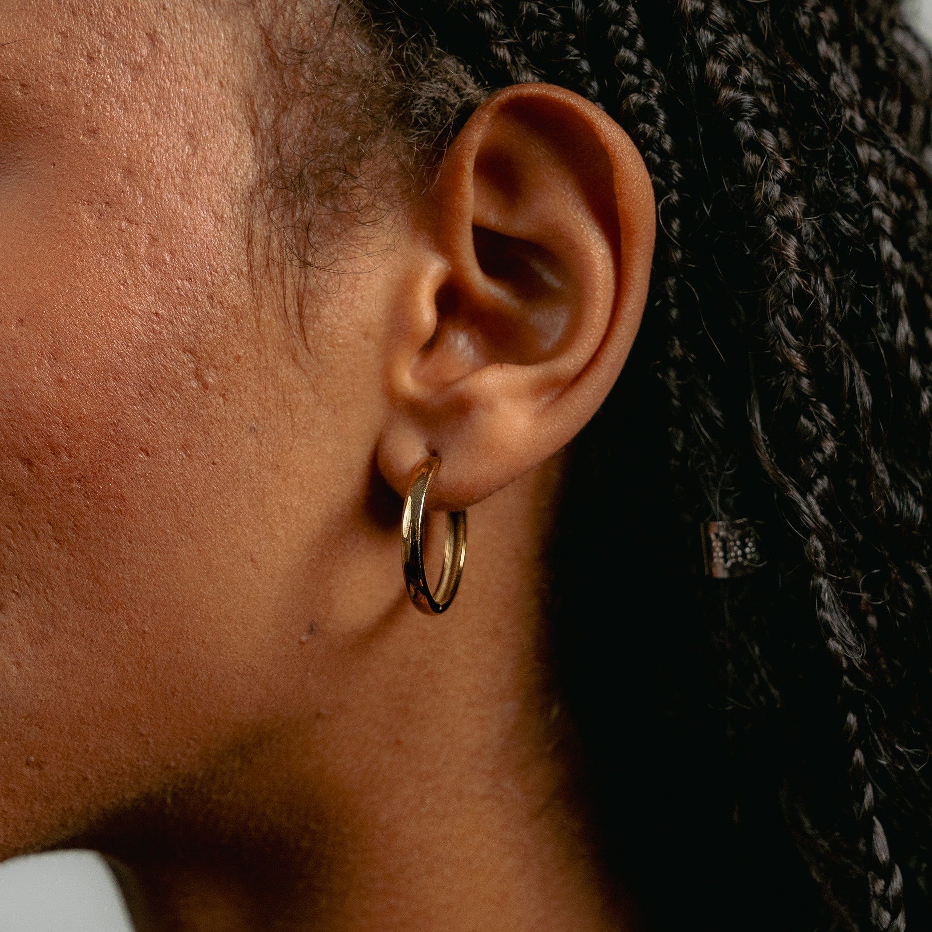Close-up of a persons ear with braided hair, wearing Glasâje Gertrude Hoops earrings (23mm, 9ct solid gold). Skin texture is visible against a neutral light background.