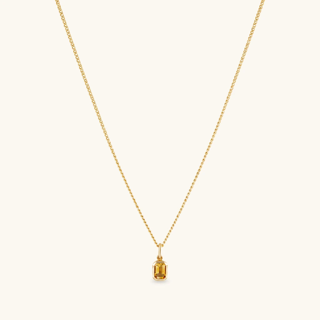 A delicate gold chain necklace with a small rectangular baguette-cut yellow gemstone pendant, displayed against a solid off-white background.
