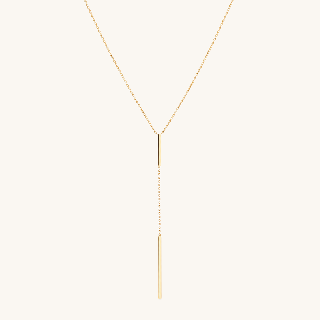 A delicate gold Y-necklace with a fine chain and a slim vertical bar pendant, displayed against a solid off-white background.