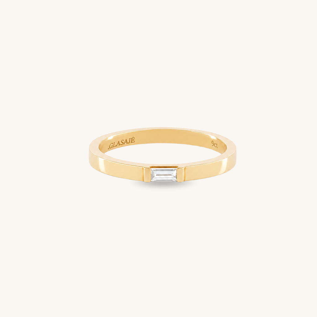 A slim yellow gold band ring with a central baguette-cut clear gemstone, shown on a clean off-white background. The brand name 'GLASAE' is engraved inside the band.
