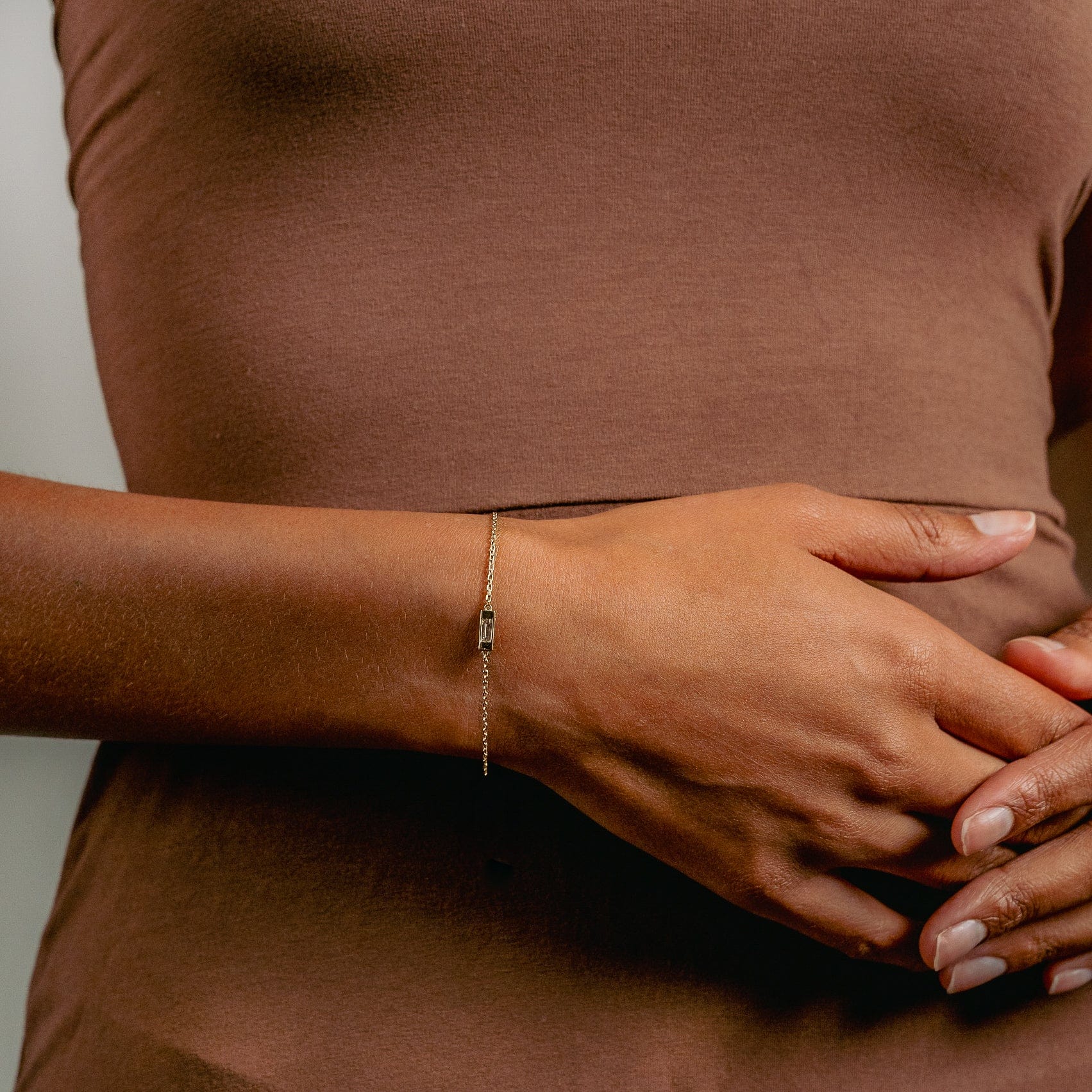 A person in a brown shirt rests one hand on their stomach, displaying the Glasâje Siglo Bracelet’s delicate solid gold link chain on their wrist against a neutral background.