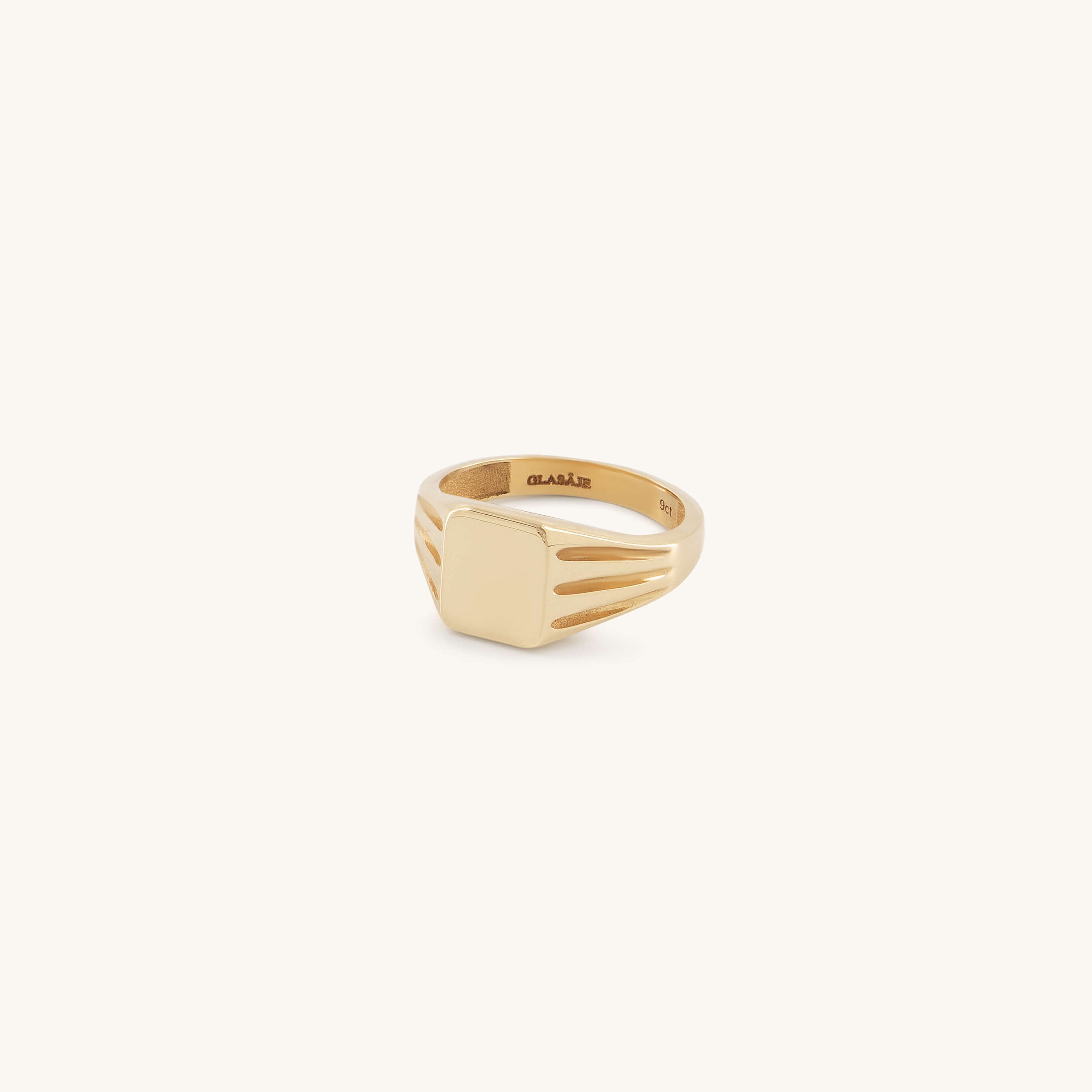 The Glasâje Signet Ring is crafted in solid gold with a rectangular flat face and debossed details, plus decorative grooves on the band, showcased against a light cream background.