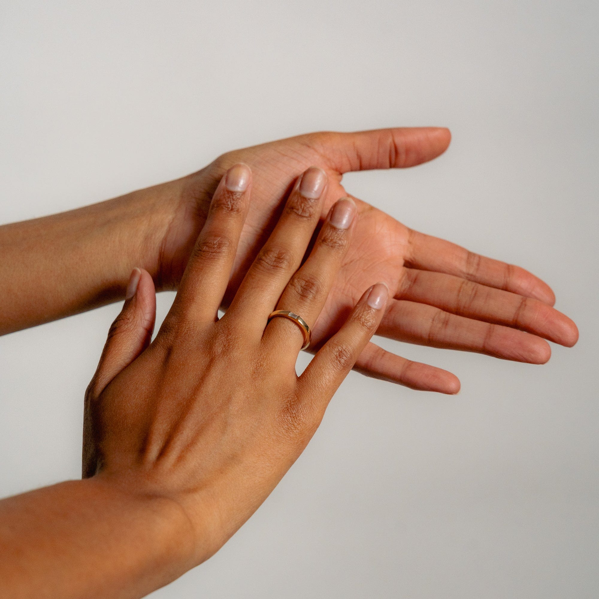 A hand with medium brown skin wears the Glasâje Siglo Ring, a simple solid gold band, while gently touching another hand’s palm against a plain light background.