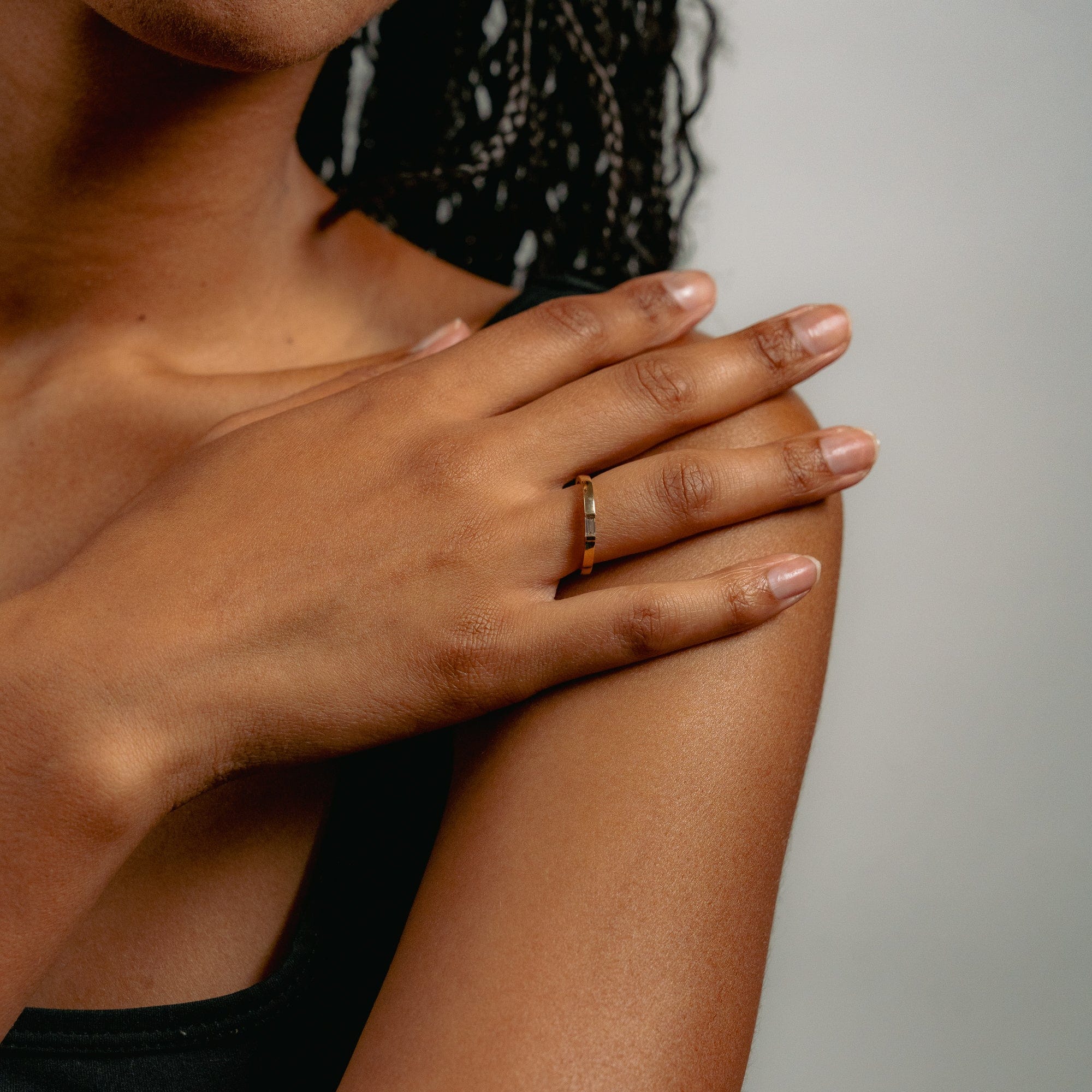 Close-up of a hand resting on a shoulder, showcasing Glasâjes Siglo Ring—an ethical diamond set in a solid gold band. The person has medium brown skin and wears a black sleeveless top against a neutral background.