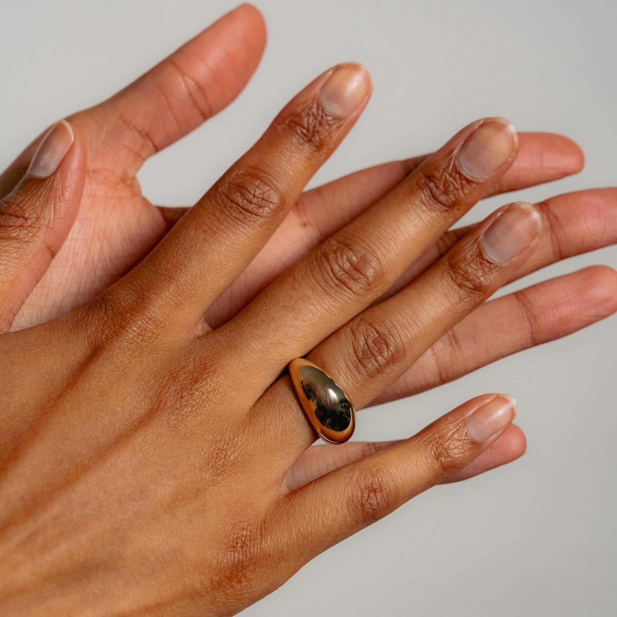 A close-up shows a hand wearing the smooth, shiny solid gold Dôme Ring by Glasâje on the middle finger, as another hand gently touches the ring finger against a plain, light background.