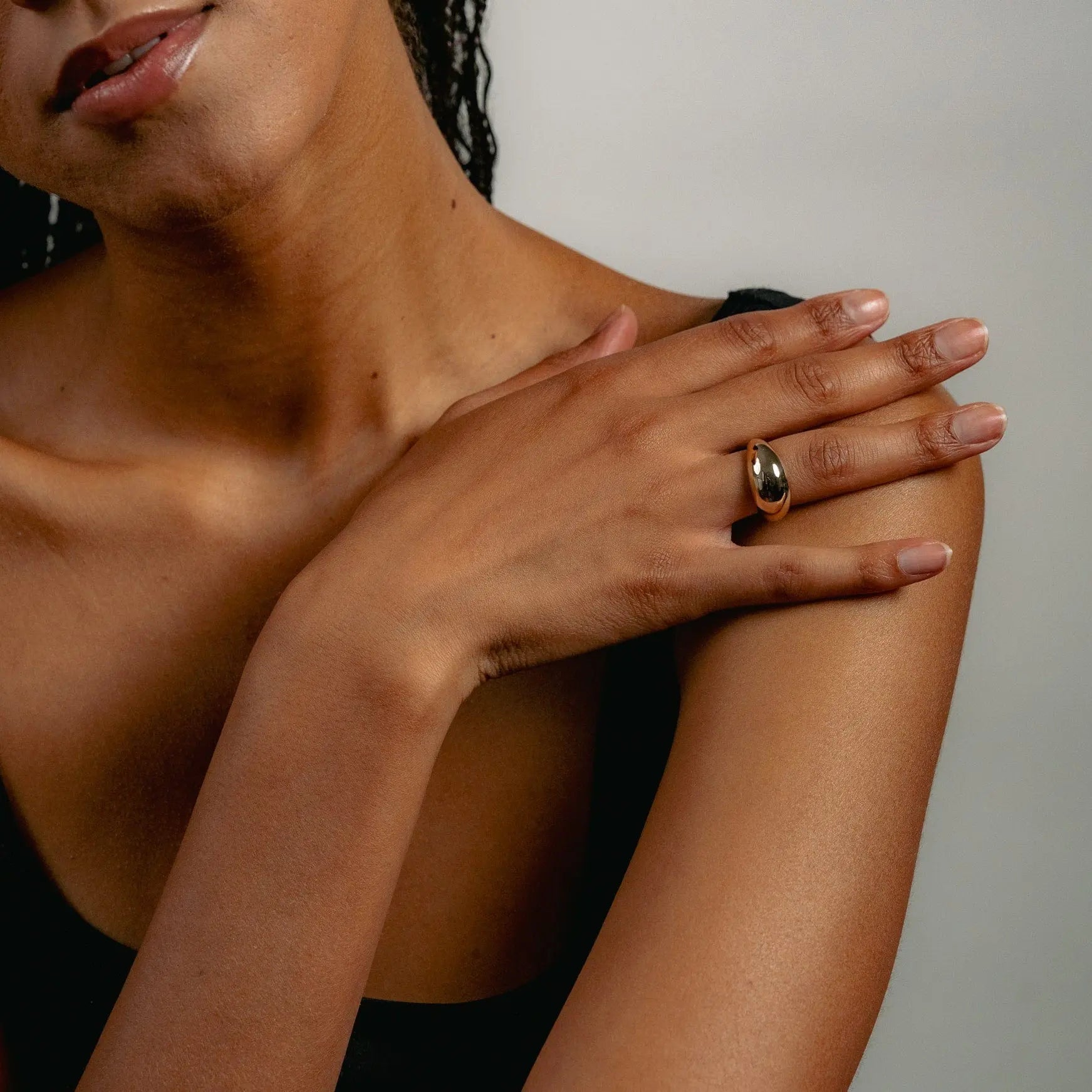 A person with braided hair rests a hand on their shoulder, wearing the solid gold Glasâje Dôme Ring. A black top and neutral background highlight the lower face and upper body, drawing attention to the ring.