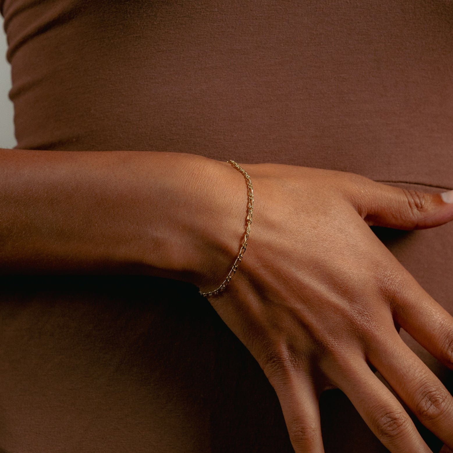 Close-up of a woman's arm wearing a delicate gold bracelet against a brown fabric background. The image conveys a sense of elegance and simplicity.
