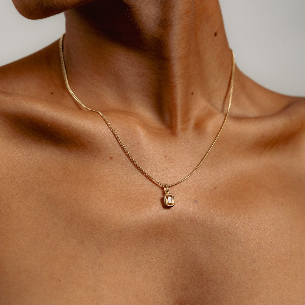 A close-up of a persons neck and upper chest wearing a thin gold chain necklace with a small, rectangular pendant. The background is plain and the persons skin tone is medium brown.