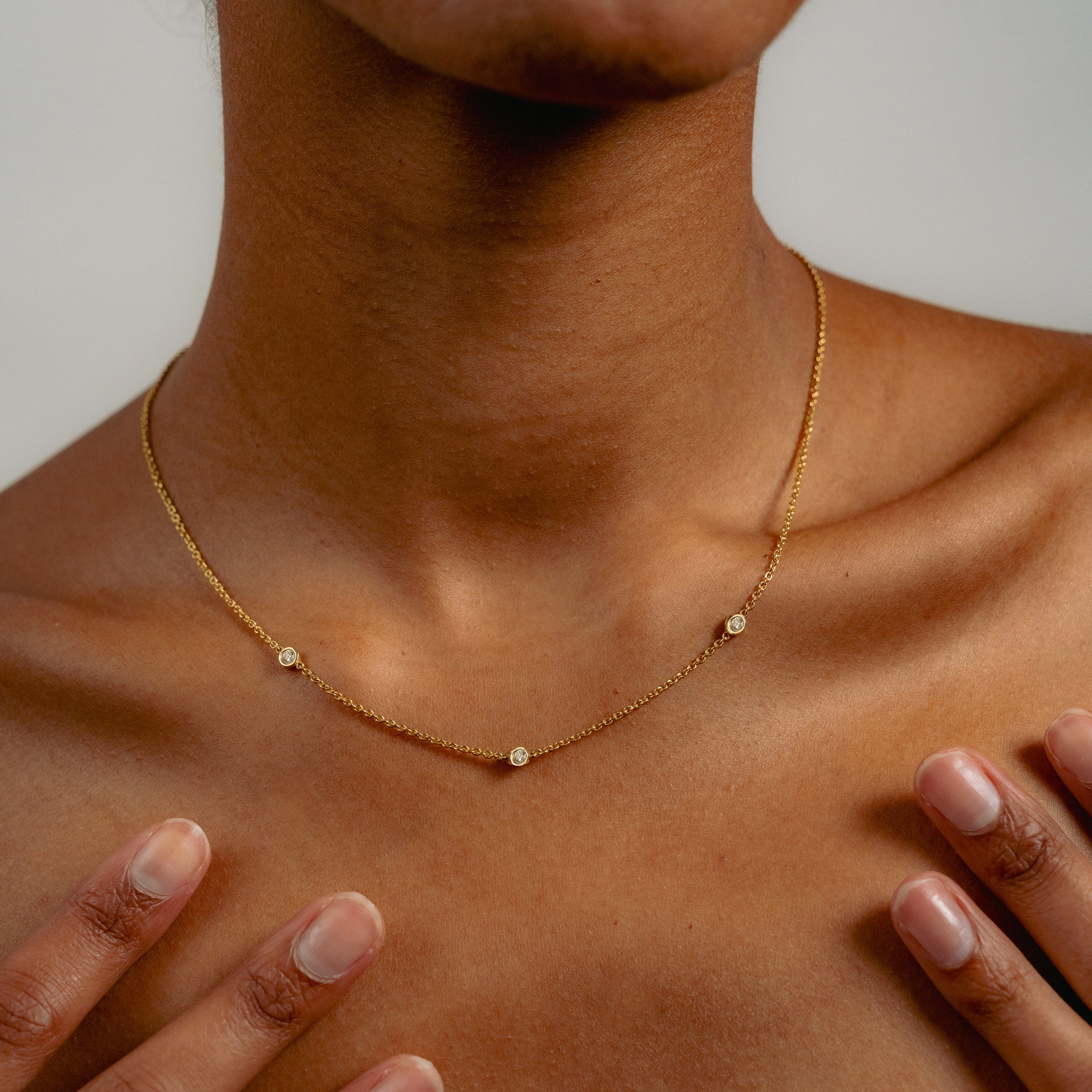 A close-up of a neck adorned with the Glasâje Satellite Necklace, crafted in 9ct solid gold and featuring three small round pendants. Manicured hands rest gently on the collarbones.