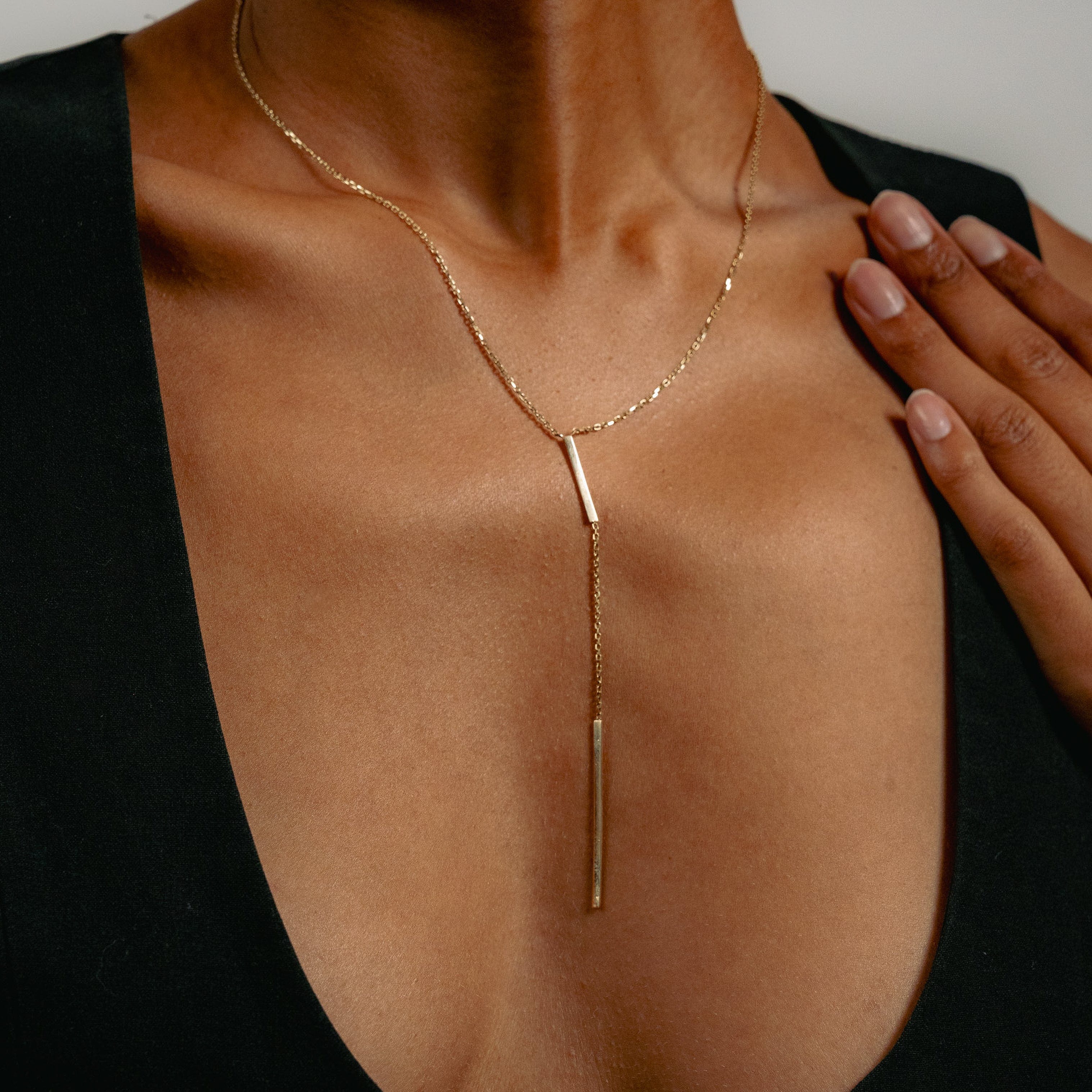 Close-up of a woman wearing a sleek gold Y-necklace with a vertical bar pendant. Her hand rests on her chest over a black v-neck dress, conveying elegance.