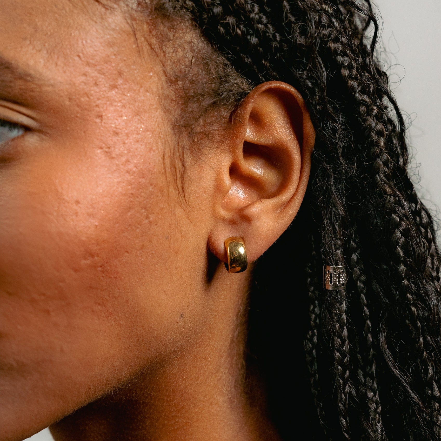 Close-up of a person with braided hair wearing Glasâje Gabriel Huggies, a timeless solid gold earring. The focus is on the ear and earring, showcasing skin texture and braids against a plain, blurred background.