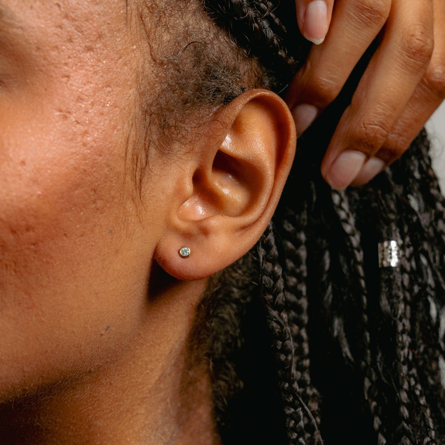 A close-up shows a person with brown skin and neatly trimmed nails wearing Glasâje Satellite Studs, their braided hair brushed aside as their hand gently touches their head.