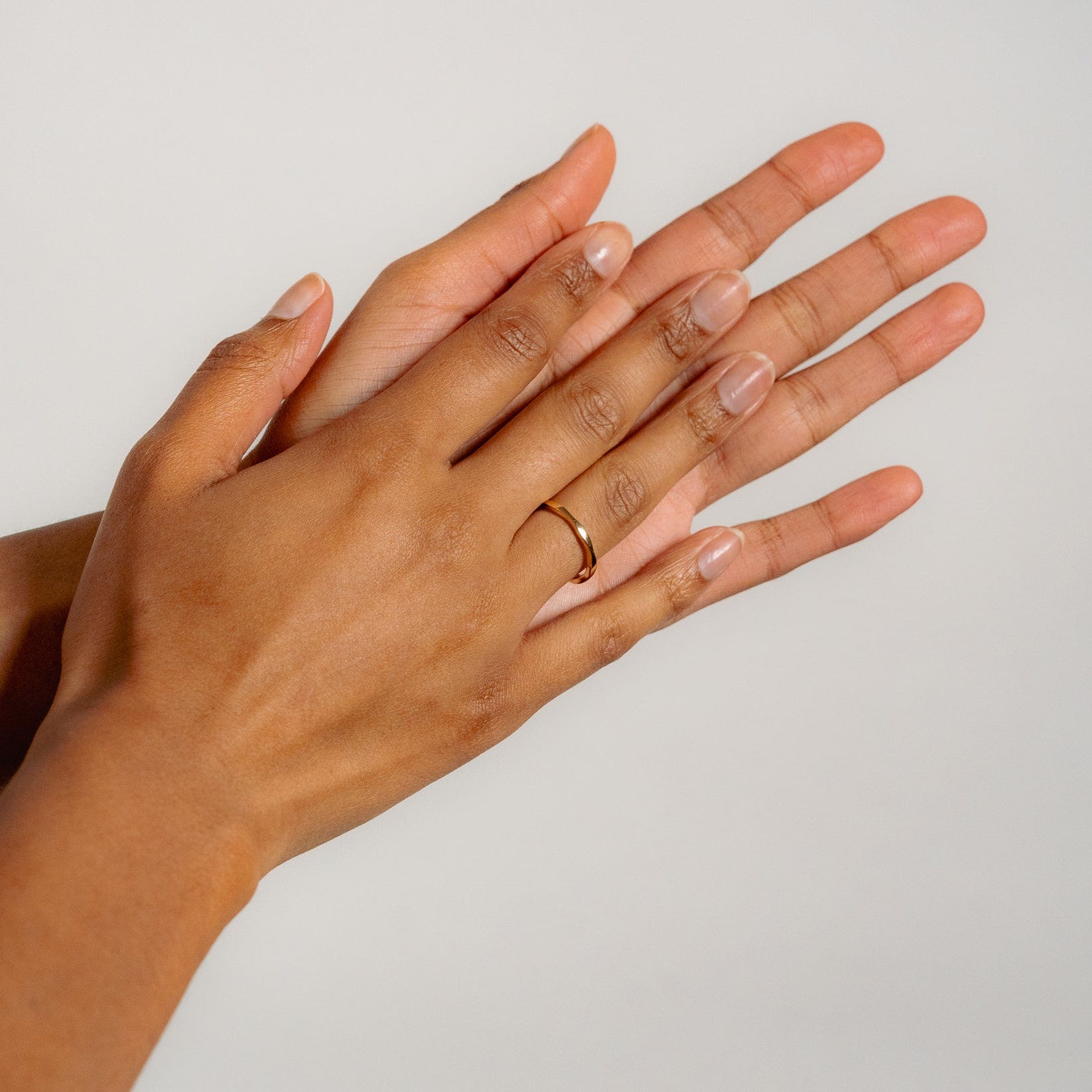 Two hands, one wearing the Glasâje Ripple Stacking Ring on the left ring finger, gently rest atop each other against a plain light background. The ring’s minimal ripple design brings effortless elegance to the serene moment.