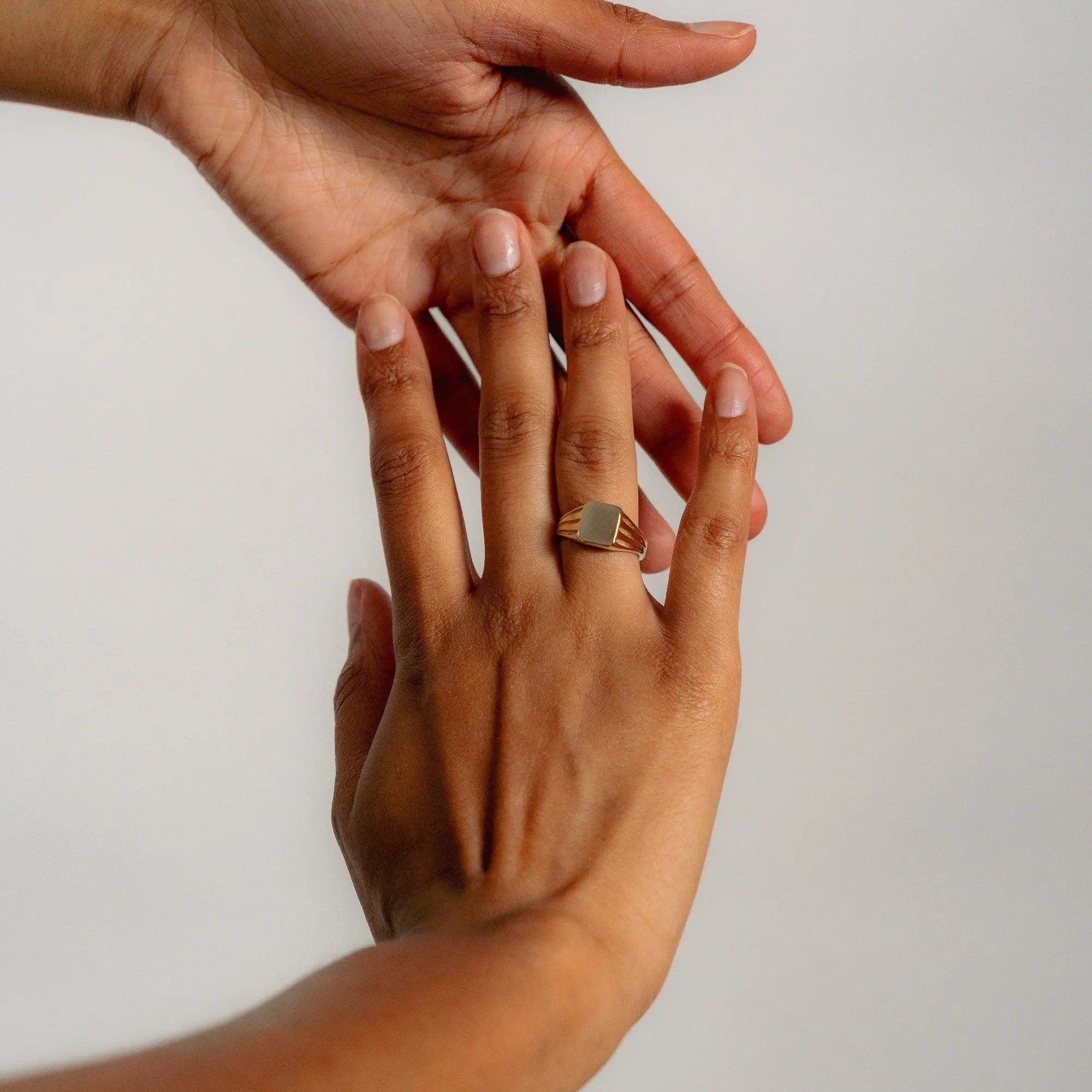 A close-up shows one hand gently touching another, which wears the Glasâje Signet Ring—a solid gold ring featuring a rectangular face with subtle debossed detailing—set against a plain, light-colored background.